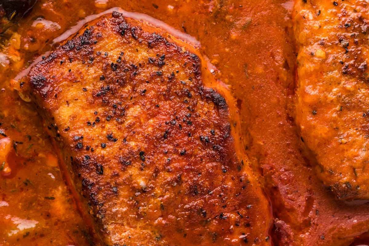 Close-up of grilled pork chops in a rich tomato sauce, showing crispy charred edges and vibrant sauce texture.