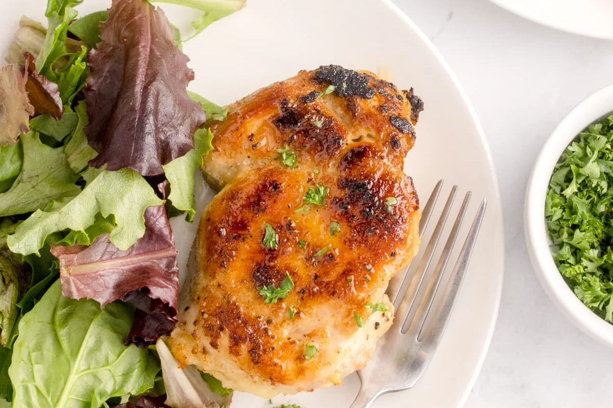 A grilled chicken breast garnished with chopped herbs sits on a white plate next to a mixed green salad. A fork is placed on the plate.