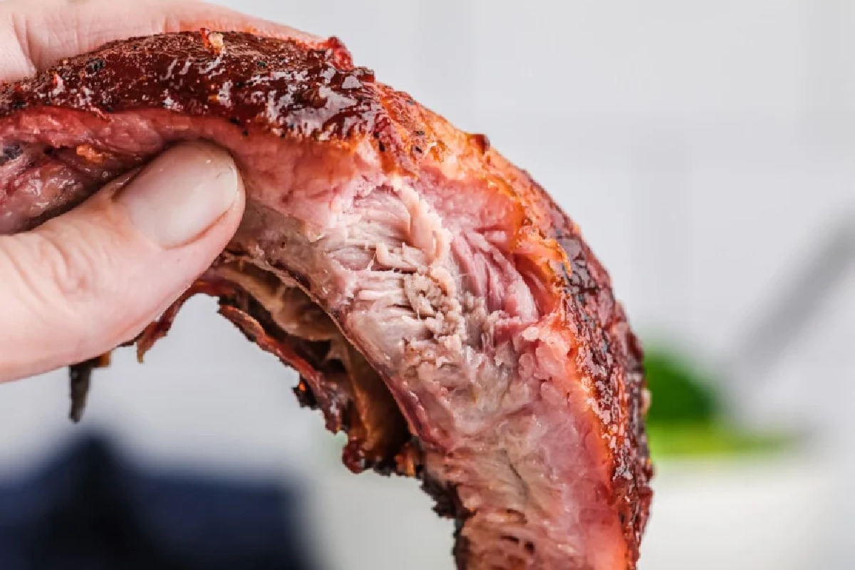 A close-up of a hand holding a barbecued rib with a shiny glaze, showing the juicy, tender meat that's sure to thrill bold taste buds.