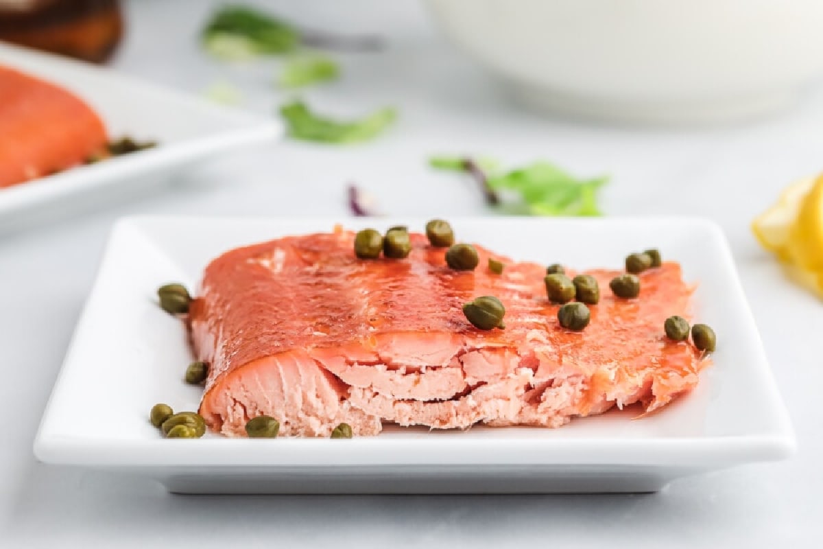 A piece of cooked salmon topped with capers is served on a square white plate.