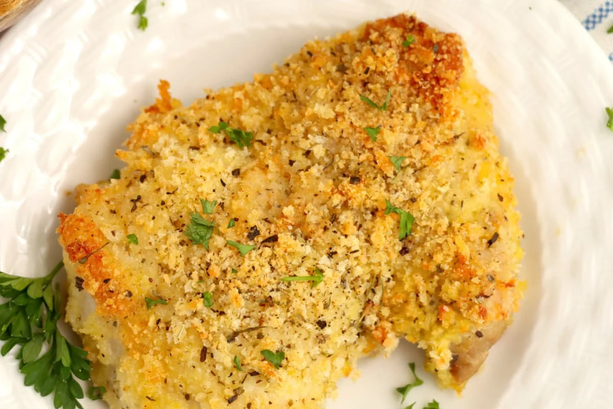 A piece of breaded and seasoned chicken or fish is served on a white plate, garnished with chopped parsley. This dish is one of those irresistible chicken recipes that everyone will love.