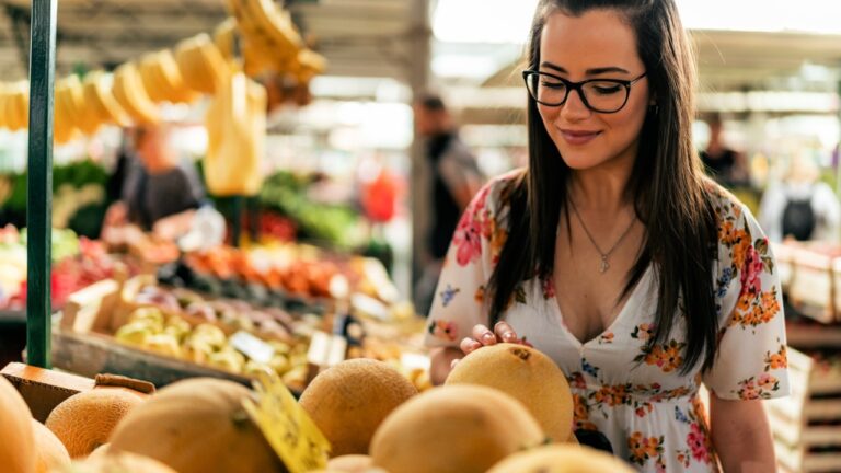A smiling girl at the market selects, taps, and smells a melon